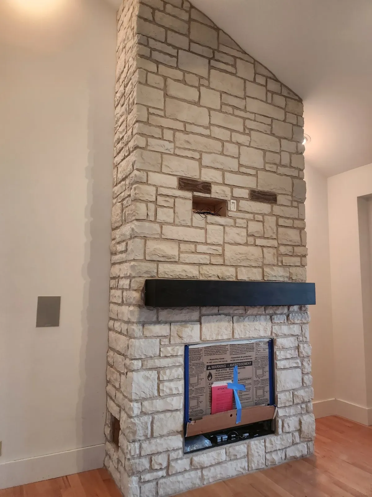 Tall light stone fireplace rising floor to ceiling with a black mantel shelf, built as an upgrade to a living room fireplace