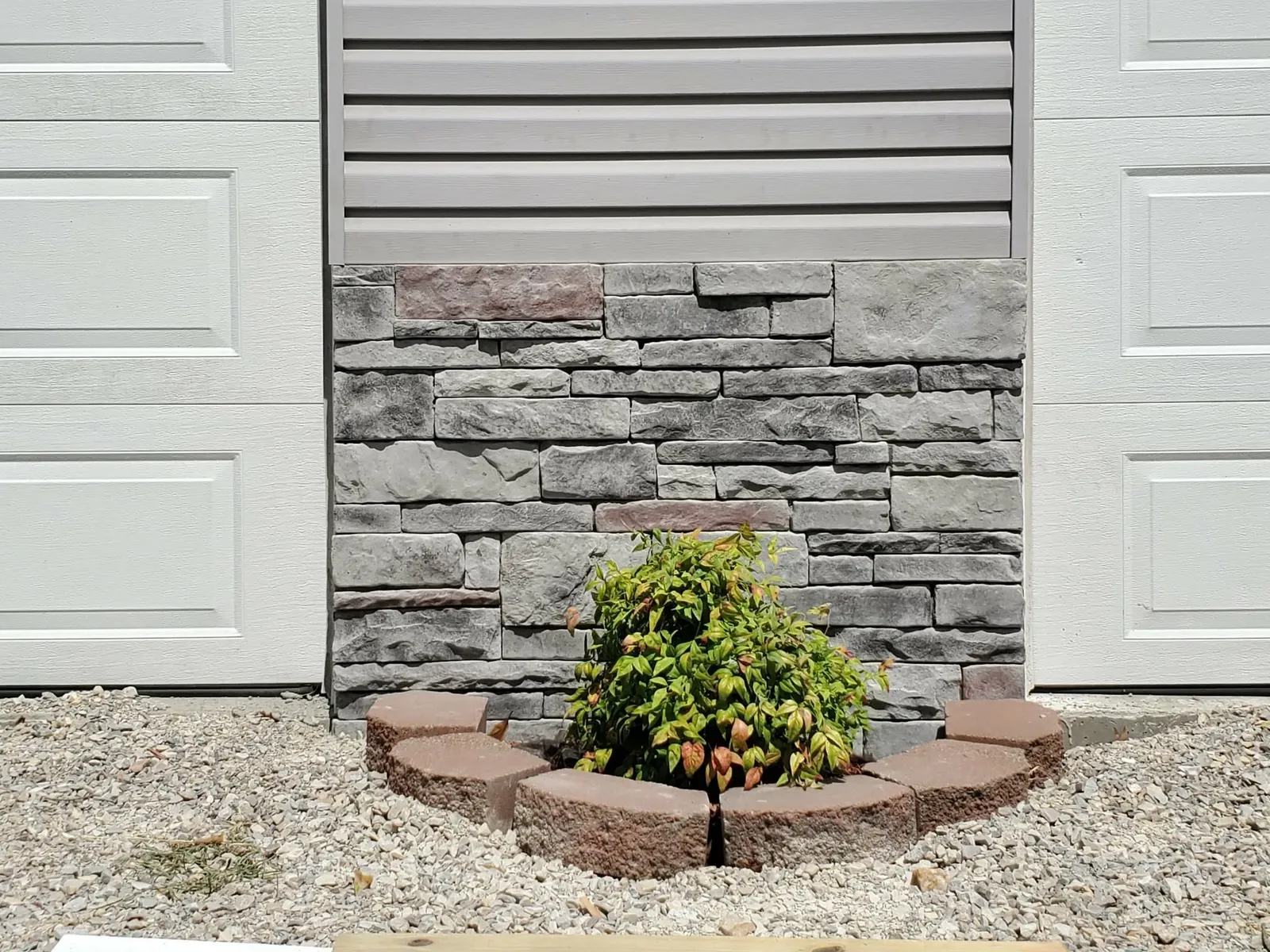 Gray stacked stone panel between two garage doors with a small landscaped bed