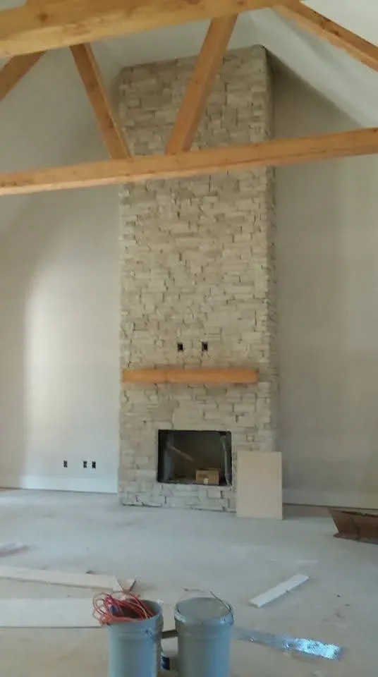Tall interior fireplace in light cream stone with a wood mantel under timber trusses, room still under construction