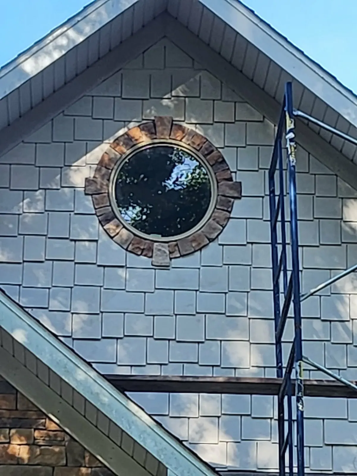 Round window in a shingled gable ringed with stone arch trim and keystones, scaffolding at the edge