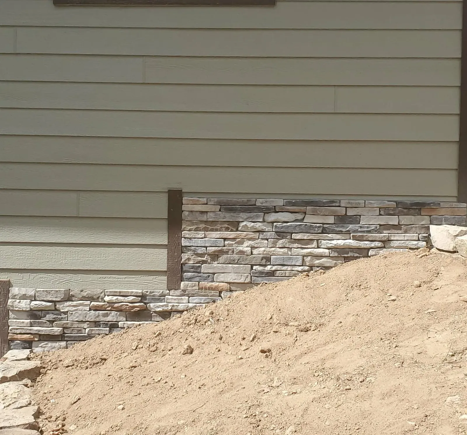 Stone wainscot stepping down a slope below house siding, with bare dirt in the foreground