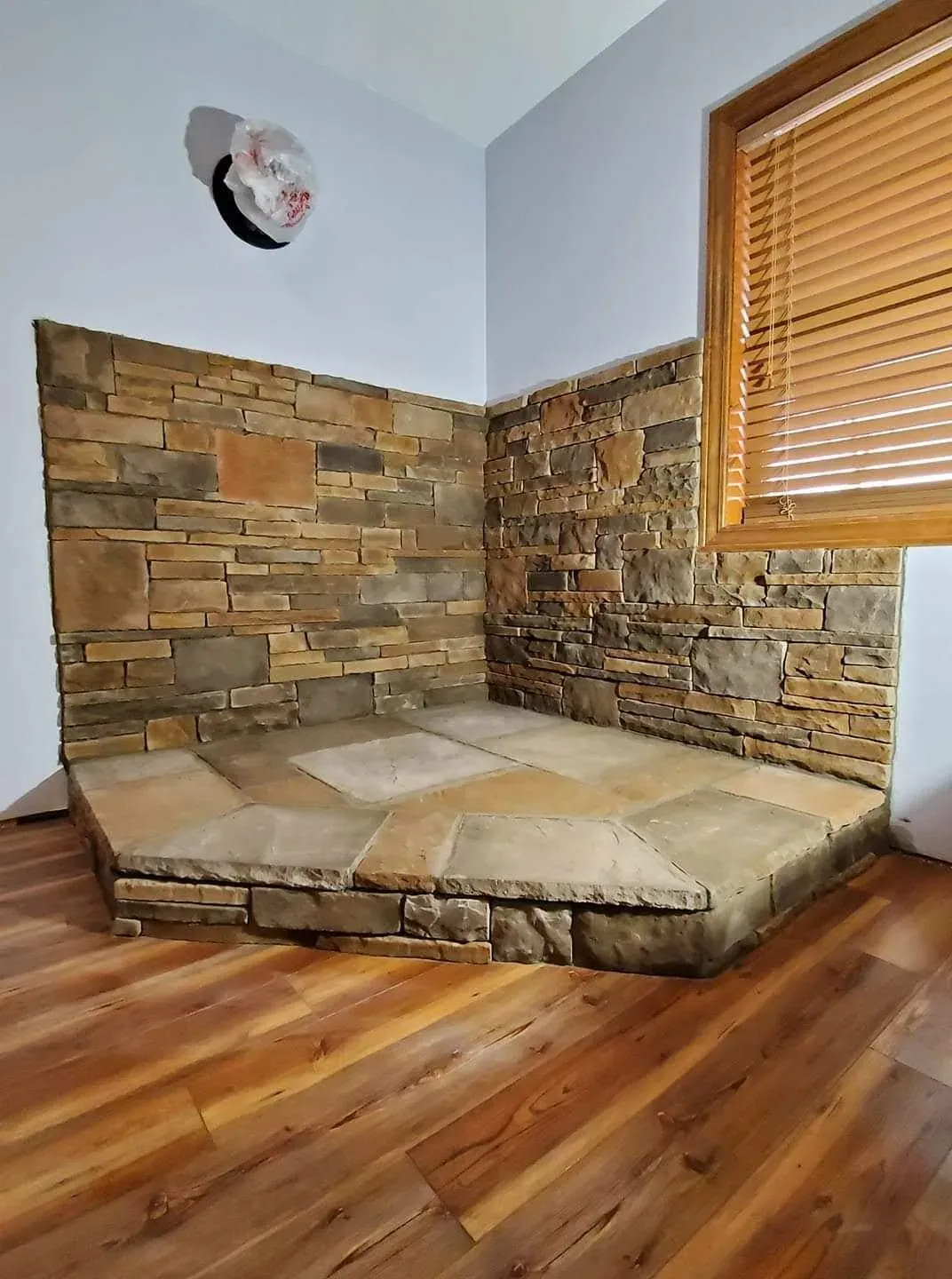 Interior stone corner with a raised stone-slab hearth platform built for a wood stove, on a wood floor